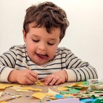 Primer plano de un niño muy concentrado realizando manualidades sobre una mesa con material y recortes de colores.