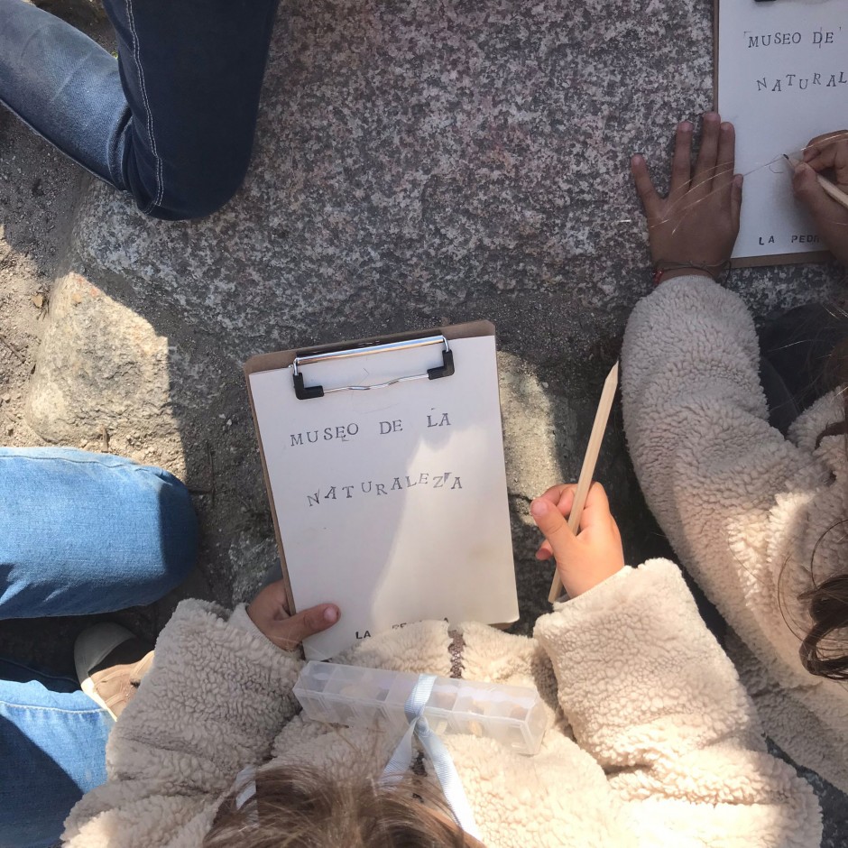 Vista superior de varias personas (menores) sentadas sobre un suelo rocoso. Dos de ellas sostienen entre sus manos lápices y portapapeles. Uno de los portapapeles tiene impreso el texto: 'Museo de la naturaleza'.