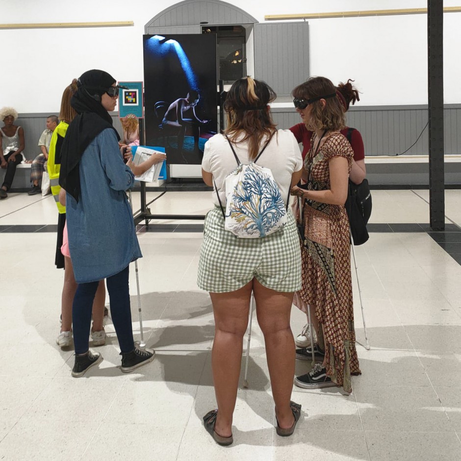 Vista general de la instalación «El sonido de la luz» en una sala espaciosa con las vigas del techo a la vista. En el centro, un grupo de seis jóvenes interactúa con la obra. Cuatro de ellas sostienen un bastón blanco, que comúnmente usan las personas con discapacidad visual. Todas llevan unas gafas, que forman parte de la experiencia de la instalación. Al fondo, una imagen a tamaño real de una figura humana sumida en penumbra, iluminada por un suave haz de luz, sentada en una silla y utilizando un teléfono, parte de la instalación.