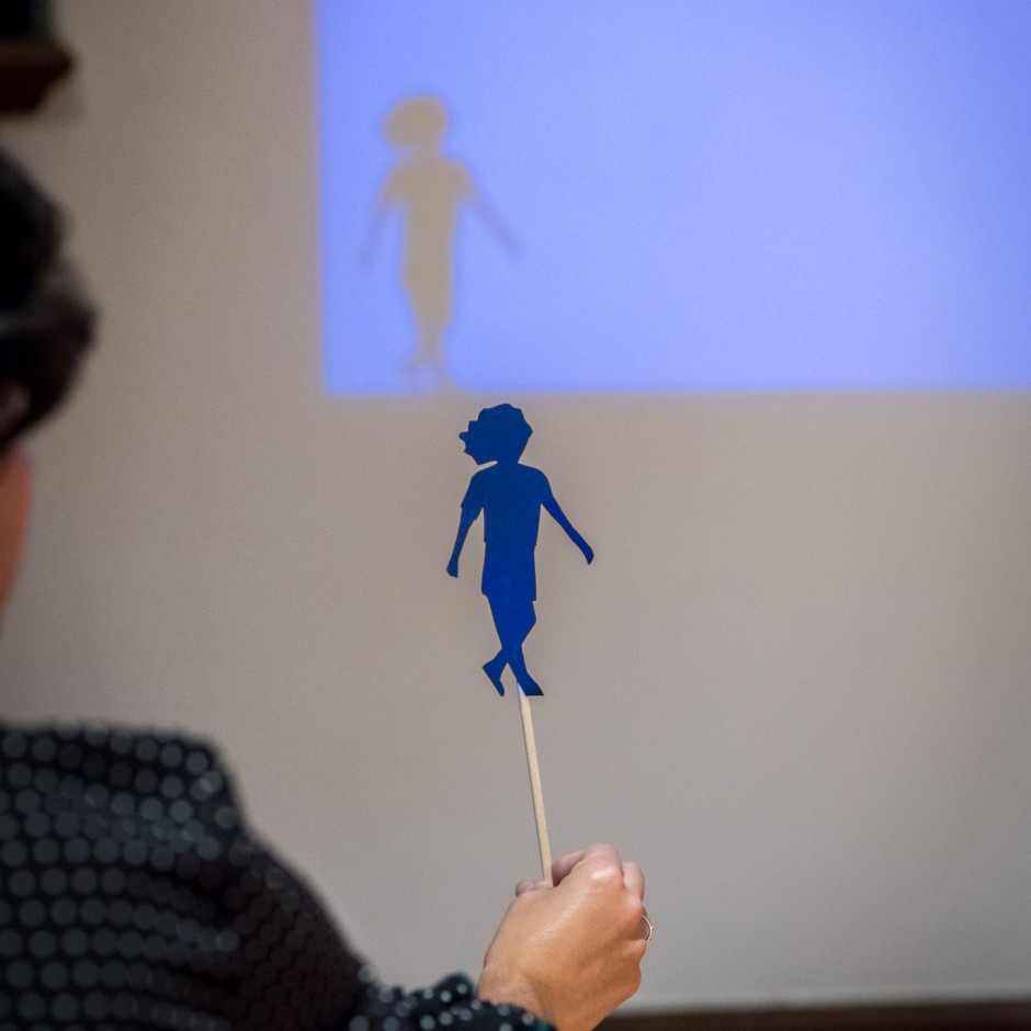 Vista posterior de una persona sosteniendo una silueta de papel de un niño que proyecta su sombra en la pared a través de la luz, creando la ilusión de que la sombra es más grande y viva.