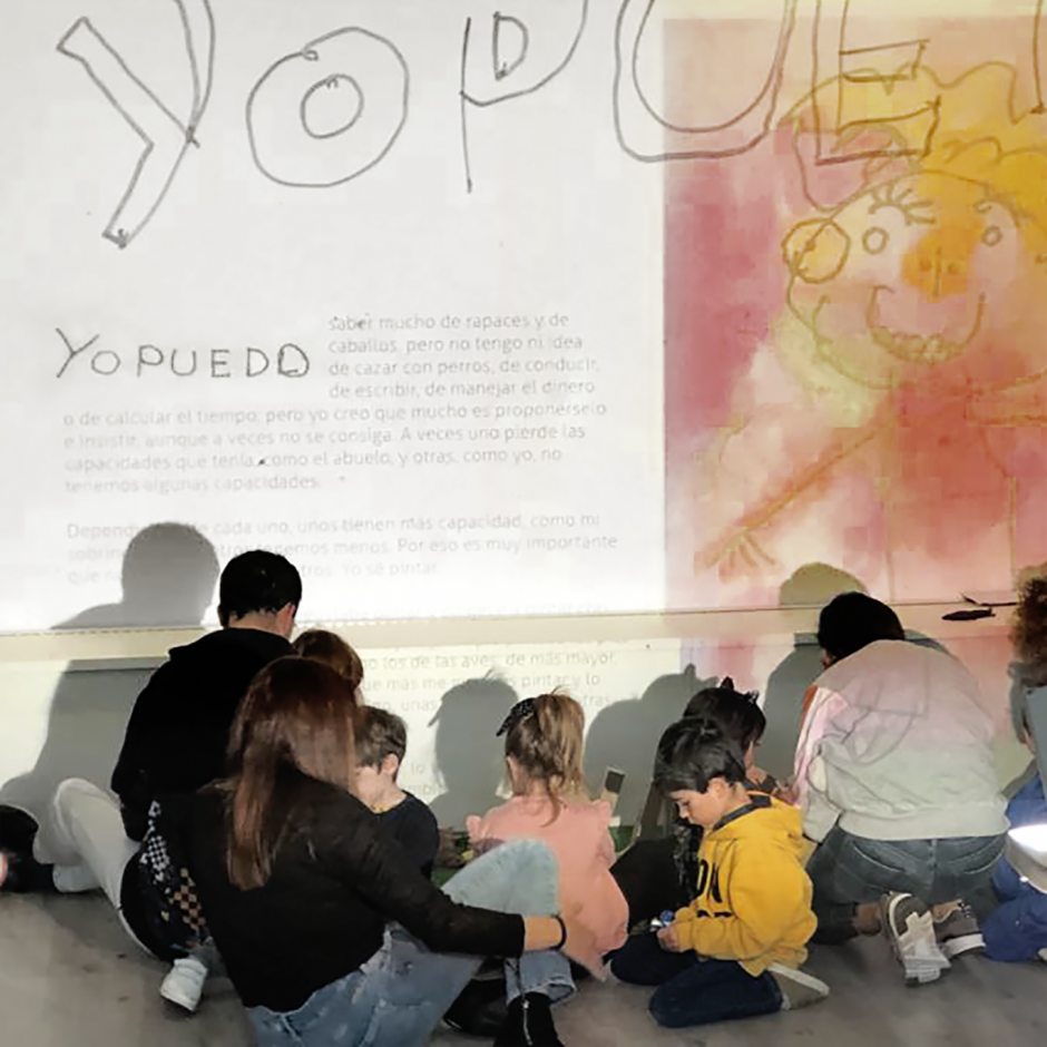 Vista posterior de un grupo de peques sentados en el suelo, frente a una gran proyección en la pared que dice "YO PUEDO" junto con un texto y una ilustración con forma humana realizada en tonos amarillos y rosados. La ilustración, realizada por Raúl, sonríe.