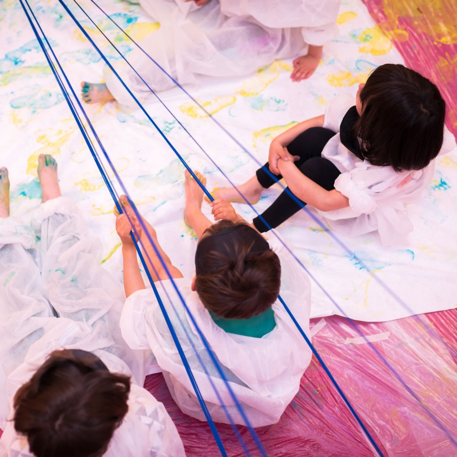 Vista superior de tres menores, con trajes de protección, sentados en el suelo con las piernas estiradas sobre una lona plástica llena de pintura.