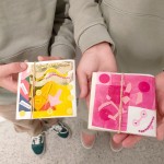 Vista detalle de dos manos que sostienen cuadernos pequeños de arte creados a mano con papel celofán en tonos amarillo y rosa, lentejuelas y recortes de papel con formas geométricas y palabras superpuestas.