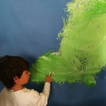 Vista posterior de un niño pintando con su manos trazos verdes sobre papel un papel azul en una Propuesta de experimentación de la obra The River.