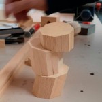 Vista detalle del taller de Manualia. Sobre una mesa de trabajo, en primer plano, tres pequeñas piezas de madera octogonales. En el fondo, herramientas de tallado y los brazos de una persona trabajando.