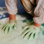 Vista superior de dos manos infantiles cubiertas de pintura verde, pintando sobre una lona azul en el suelo.