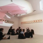 Un grupo de niños sentado en el suelo de una sala observa formas geométricas en forma de láminas proyectadas en el techo.