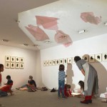 En una sala con dibujos en papel expuestos en pared, un grupo de niños observa las formas que proyectan en el techo los recortes de celofán rojos y realiza manualidades.