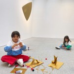 Dos niñas sentadas en el suelo, junto a la obra de latón Mask, haciendo manualidades con cartulinas de color dorado.
