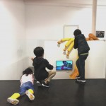 Vista posterior de tres niños contemplando la obra 'El Fliscorno y el Zapato de Gamuza' (cuero sintético + instalación vídeo).
