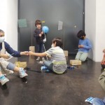 Vista frontal de un grupo de niños junto a una organizadora creando, en el suelo, una copia de la obra 'Biruta’ de Carlos Nunes con palitos, papel de celofán y globos azules. 