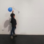 Vista perfil de un niño mostrando a cámara su creación. Una veleta en forma de cruz realizada con palitos de madera, papel de celofán y globos en los extremos del travesaño.