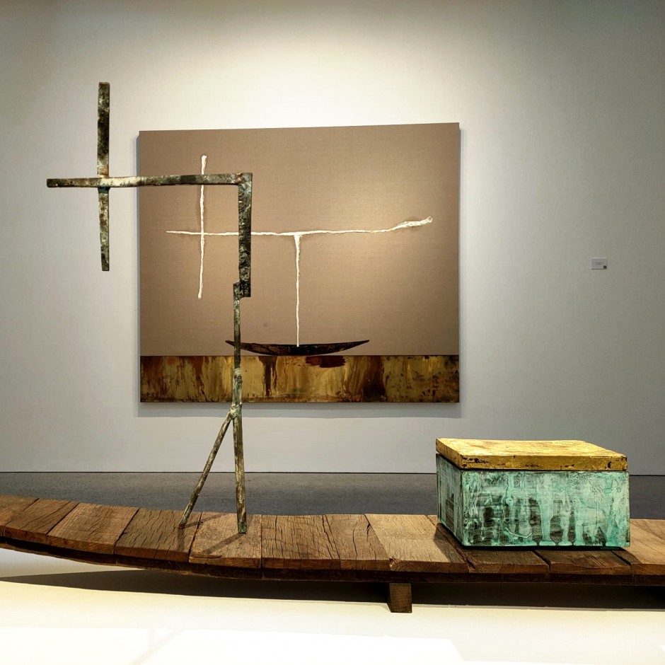 Sala de exposición. Al fondo, la obra 'Avión blanco sobre barca y mar’ sobre lienzo en hierro, cobre y collage. En primer plano, la escultura 'Barca con cofre' en madera, bronca y pan de oro.