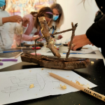 Detalle de una recreación de la obra ‘Barca con cofre’ en una mesa junto a un dibujo a lápiz. Tras ella, el grupo de menores y acompañantes haciendo manualidades.