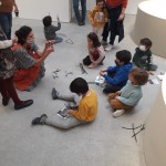 Un grupo de niños y niñas realizando manualidades en el suelo con María y familiares junto a obras en pequeño formato.