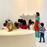 Instantánea de María explicando a un grupo de niños y niñas las características de tres obras de pequeño formato realizadas en bronce dorado. Entre ellas, «Henchida».
