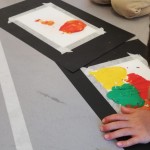 Detalle del grupo infantil creando en el suelo una obra con cartulinas y pintura roja, amarilla, naranja y verde.