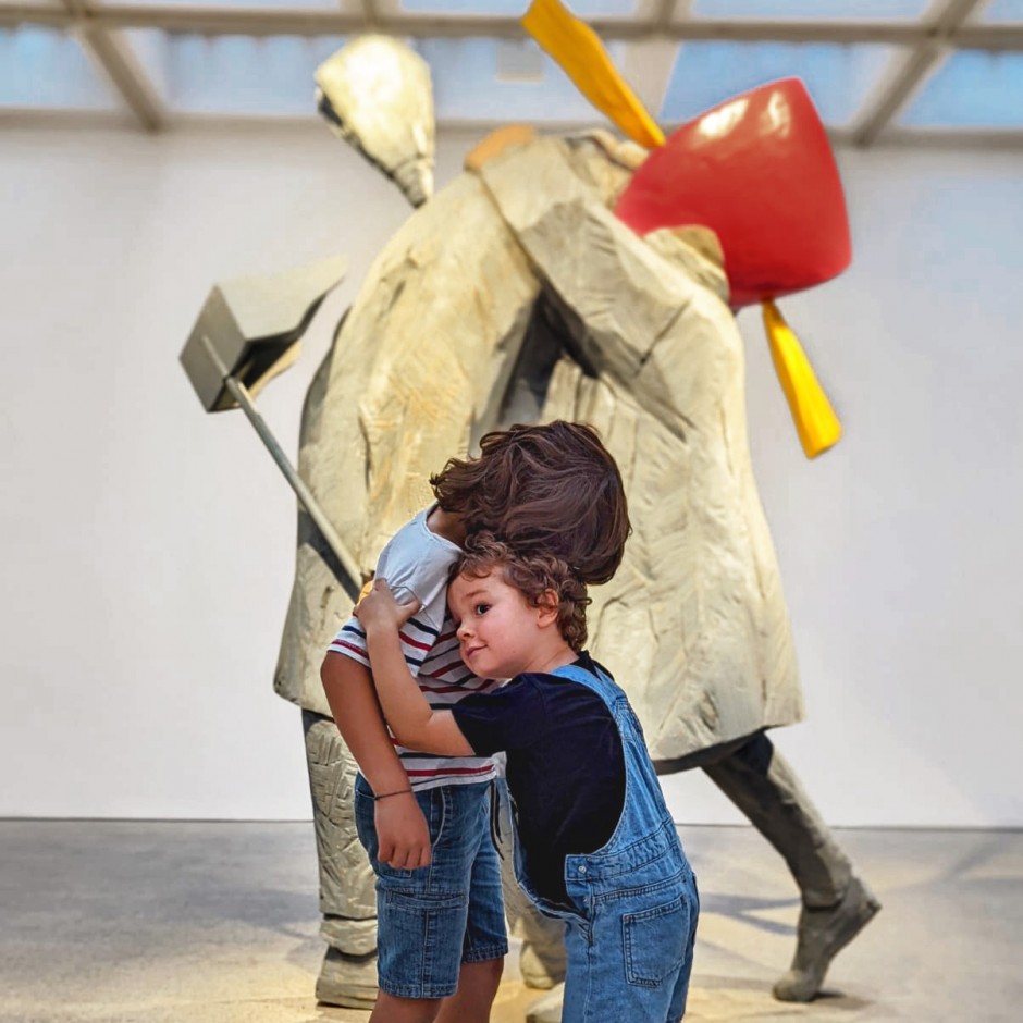 Vista frontal de la escultura en madera 'A filla da porteira', formada por dos figuras abrazadas. Una de ellas, lleva una escoba y un recogedor. Frente a la escultura, dos hermanos que asisten al taller reproducen el abrazo. 