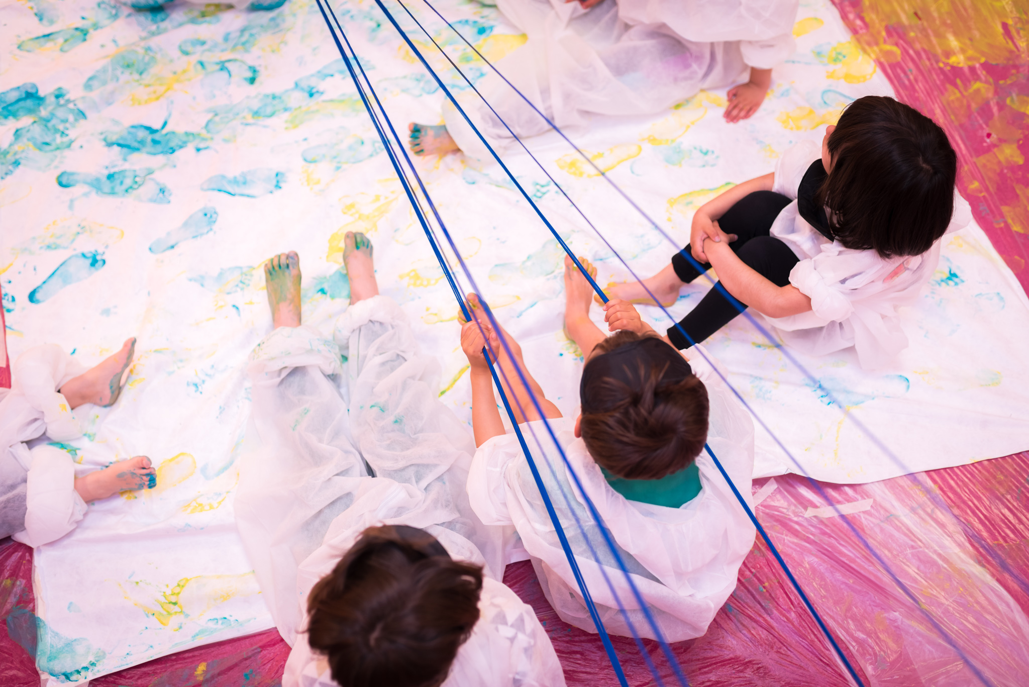 Vista superior de cuatro niñas y niños sentados sobre una tela pintada, tensando cuerdas sobre esta.