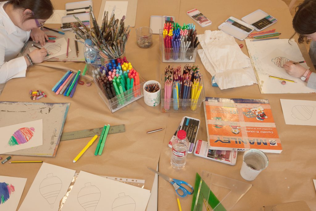 Instantánea del taller “Pintapeonzas: Down Madrid”. [Descripción de imagen] Dos niñas pintan sobre una mesa con material de manualidades: lápices, cuadernos y hojas, entre otros.