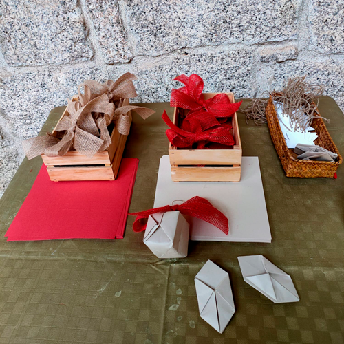 Sobre una mesa, material de manualidades: papel y lazos marrones claro, rojos; hilos; y tarjetas.