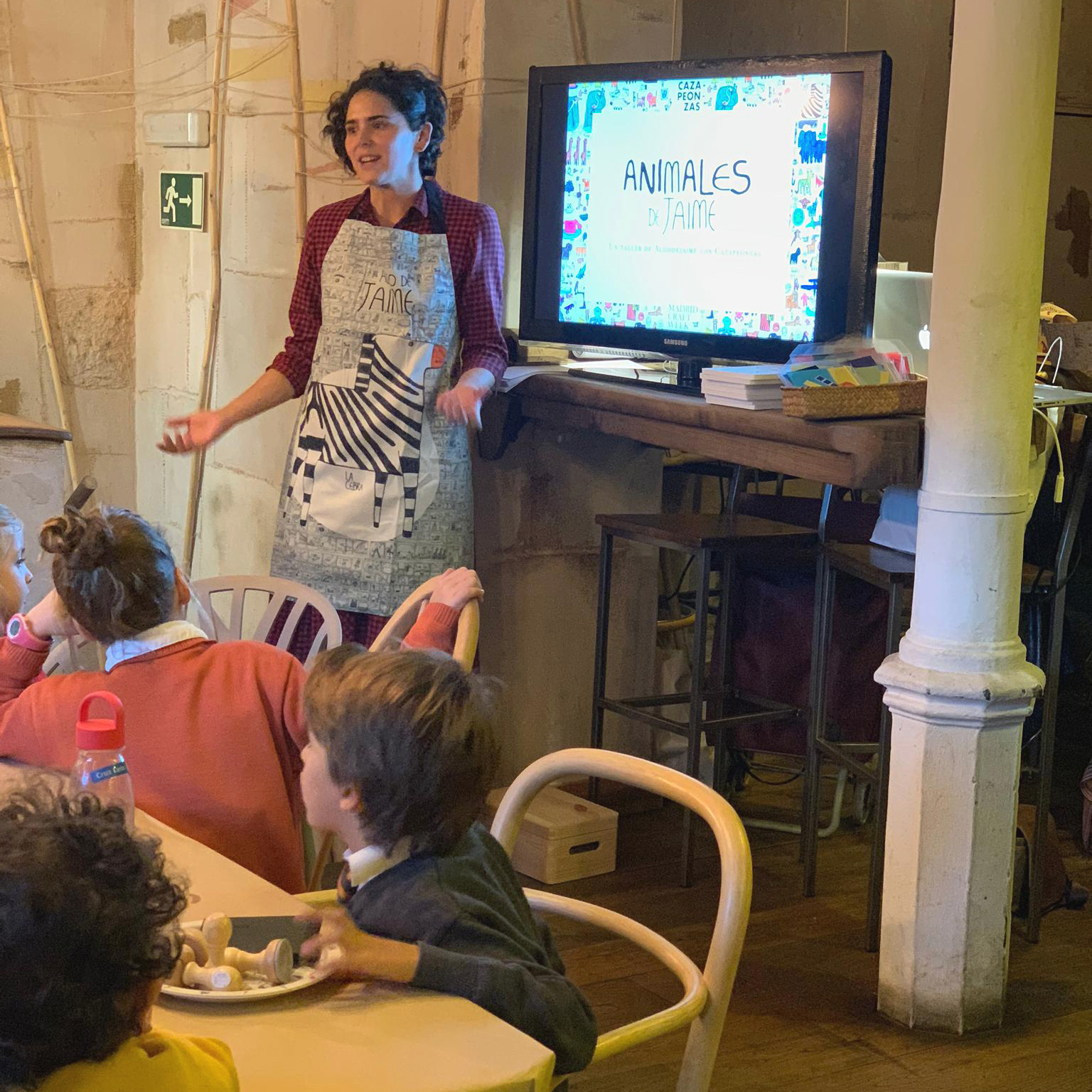 Vista lateral de María dirigiéndose al grupo reunido alrededor de mesas con una presentación en vídeo de «Animales, de Jaime».