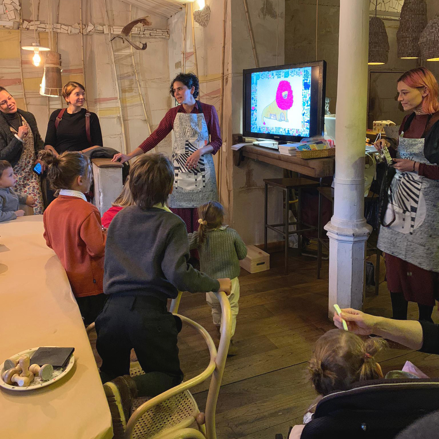 Vista lateral de María dirigiéndose al grupo reunido alrededor de mesas con una presentación en vídeo de «Animales, de Jaime». La apertura de plano abarca algunas personas más del grupo.