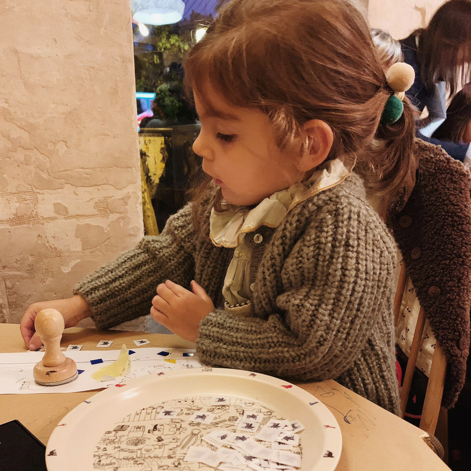 Primer plano de una niña realizando manualidades con pequeños recortes de papel, que almacena en un plato con ilustraciones de animales, y un sello de goma.