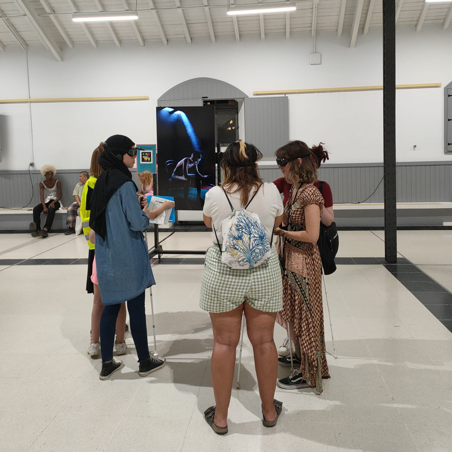 Vista general de la instalación «El sonido de la luz» en una sala espaciosa con las vigas del techo a la vista. En el centro, un grupo de seis jóvenes interactúa con la obra. Cuatro de ellas sostienen un bastón blanco, que comúnmente usan las personas con discapacidad visual. Todas llevan unas gafas, que forman parte de la experiencia de la instalación. Al fondo, una imagen a tamaño real de una figura humana sumida en penumbra, iluminada por un suave haz de luz, sentada en una silla y utilizando un teléfono, parte de la instalación.