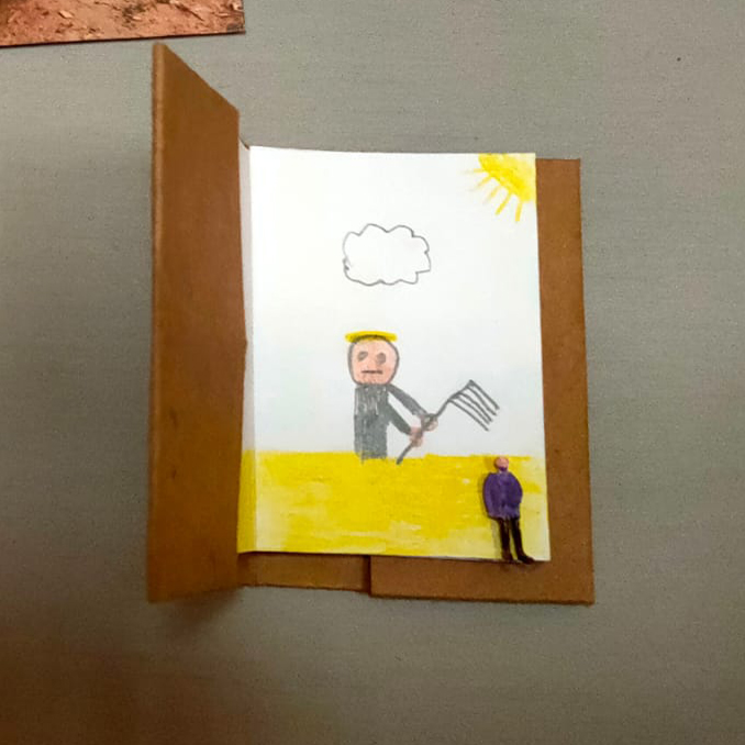 Plano detalle de una carpeta pequeña de cartón abierta con una hoja en la que hay dibujada una figura labrando la tierra bajo el sol y una nube. Sobre el dibujo, una pequeña figurita en forma de persona.