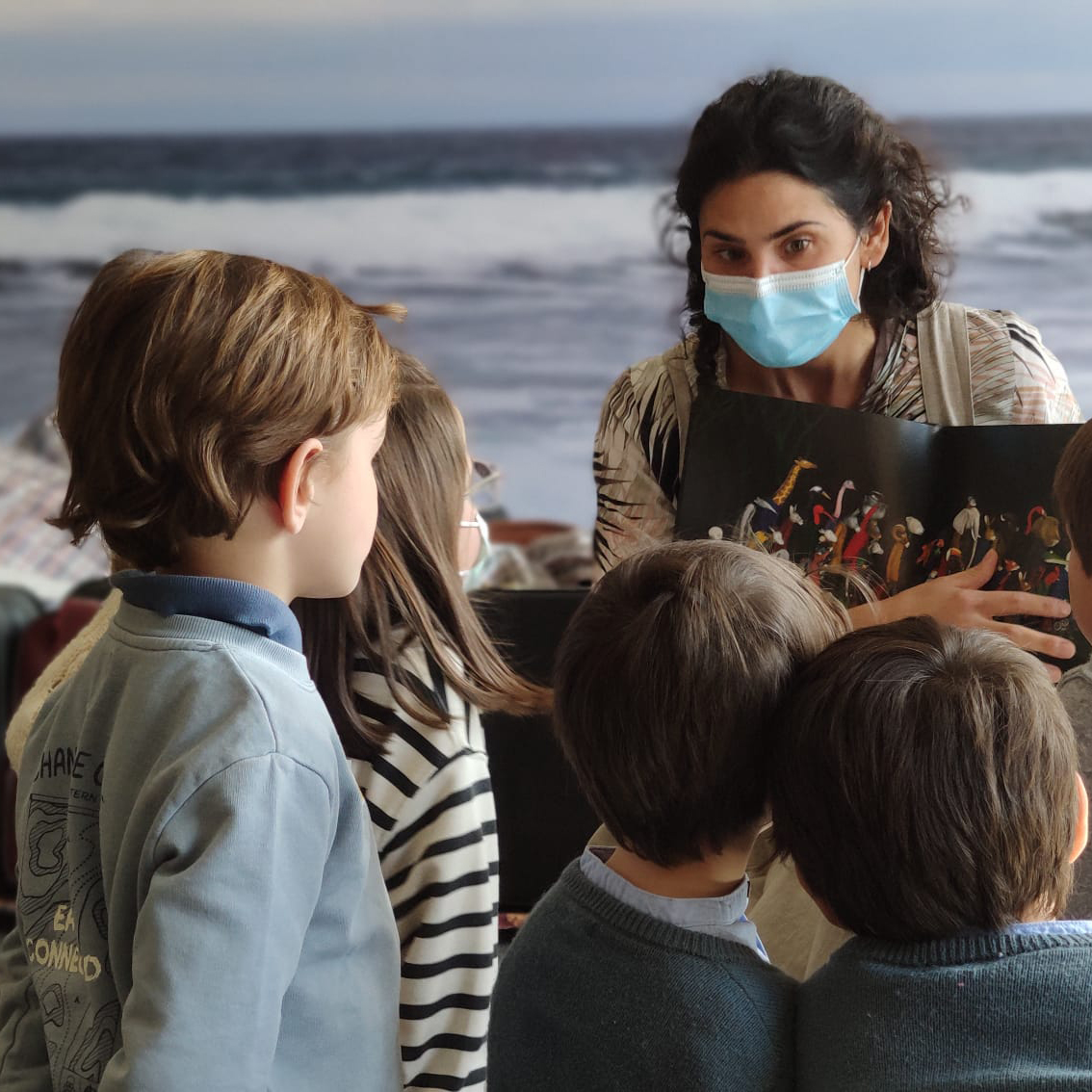 En primer plano, un grupo de niños presta atención a María, que parece contarles una historia mientras les muestra un libro abierto de par en par con ilustraciones de figuras animales. Al fondo, una superficie que simula el mar y el cielo con con tonalidades azules y violetas.