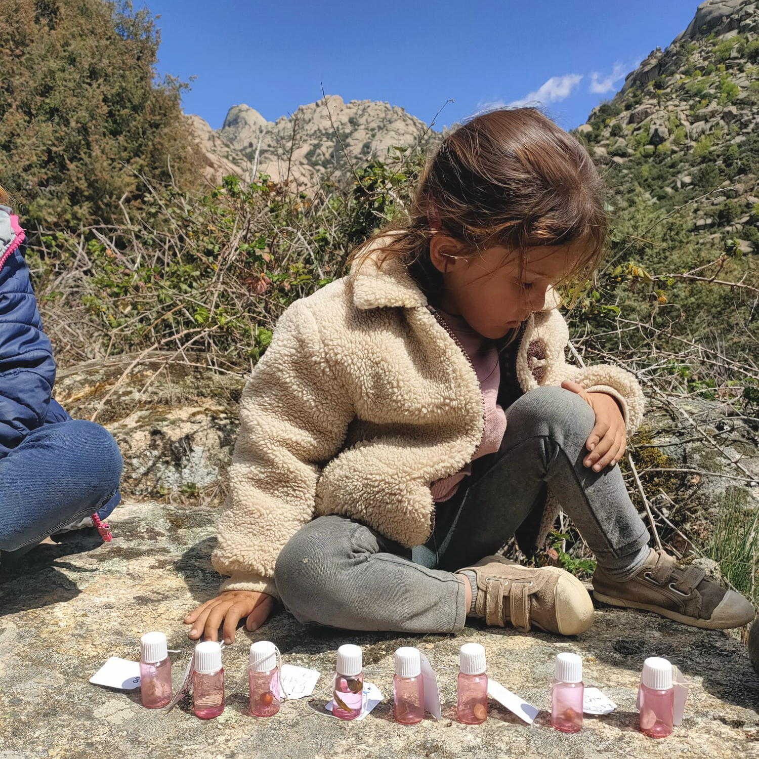 Vista frontal de una menor sentada en una roca. Frente a ella, alineadas, pequeñas botellas con piedras, rocas o minerales dentro.