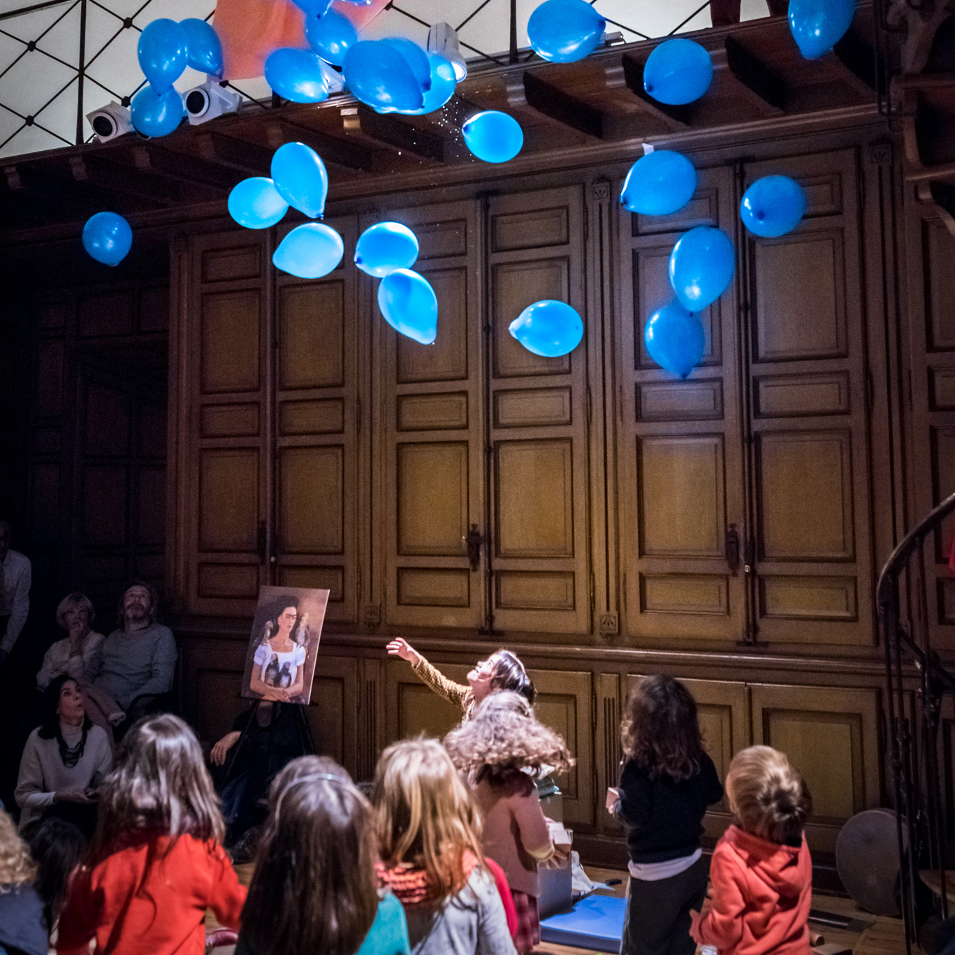 Vista lateral de María y Sara en el centro de un círculo de niños con globos azules flotando en el aire.