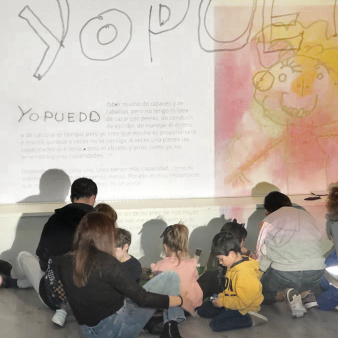 Vista posterior de un grupo de peques sentados en el suelo, frente a una gran proyección en la pared que dice "YO PUEDO" junto con un texto y una ilustración con forma humana realizada en tonos amarillos y rosados. La ilustración, realizada por Raúl, sonríe.