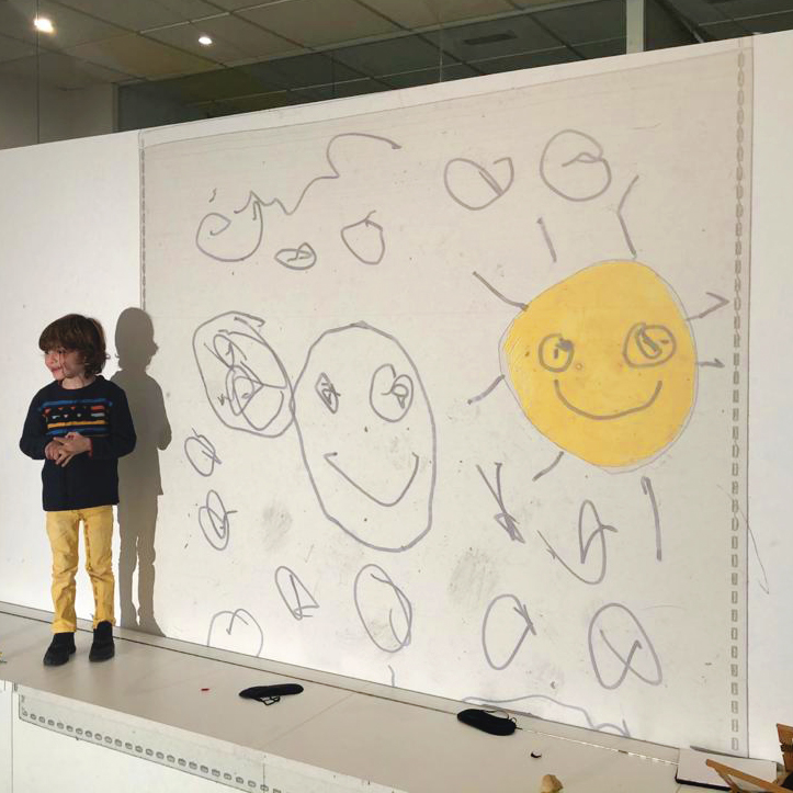 Vista lateral de un niño sonriendo frente a una pared en la que se proyectan dibujos geométricos y dos grandes soles con rostro humano. Ambos sonríen y uno de ellos está pintado en amarillo. El resto de dibujos son siluetas.