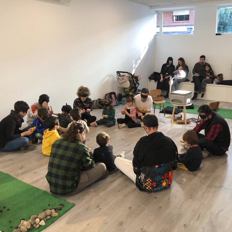 Vista general de un aula con un grupo de personas adultas y menores sentadas en el suelo y en un banco apoyado en la pared. Las personas sentada en el suelo, que es el grupo más grande, lleva antifaces para dormir.