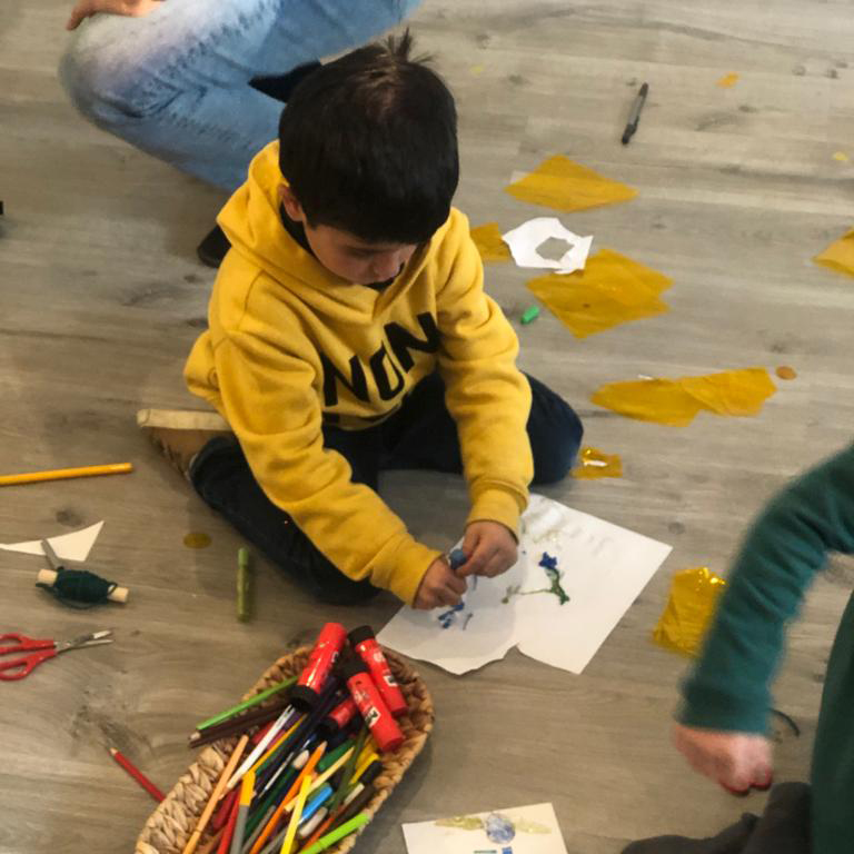 Vista superior de un niño concentrado en colorear un papel en el suelo, rodeado de rotuladores, recortes de papel celofán amarillo y tijeras.