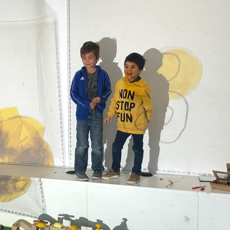 Vista lateral de dos niños frente a la pared blanca en la que se proyecta la obra de Raúl. Uno viste una chaqueta azul; el otro un suéter amarillo con la frase «Non stop stop» («Diversión sin fin»).
