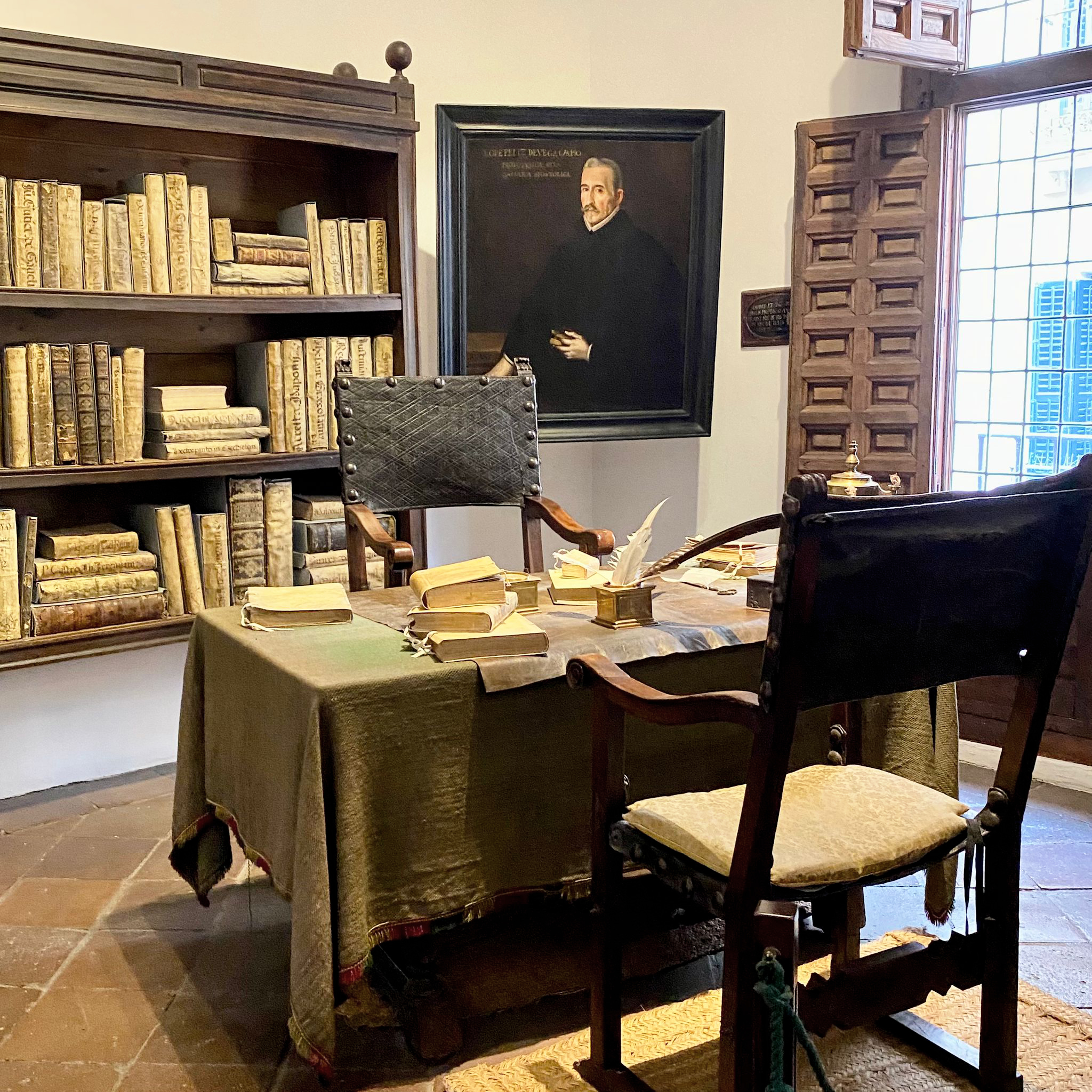 Estancia interior de la Casa Museo Lope de Vega. En ella, un escritorio con manuscritos y una pluma, dos sillas antiguas, una estantería con libros y un retrato de Lope de Vega.
