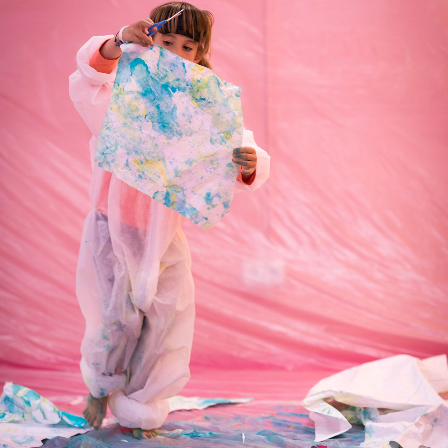Vista frontal de una niña de pie, sosteniendo una hoja de papel grande que ha pintado. Lleva un traje protector y parece mostrar su obra. Tanto la pared como el suelo están cubiertos de un plástico en tonos rosas.