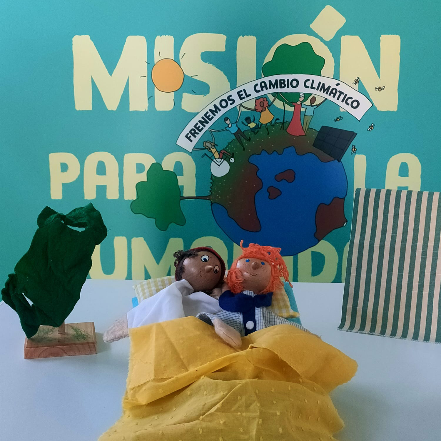 Vista detalle. En primer plano, dos muñecos de trapo con figuras humanas que parecen dormir en una cama y un árbol verde a su lado. Tras ellas, el cartel de la exposición «Misión para la humanidad», «Frenemos el cambio climático».