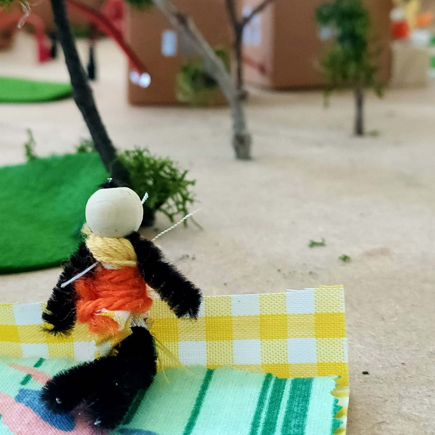 Vista detalle. En primer plano, un muñeco de trapo con figura humana sentado en el suelo sobre una manta de picnic. A su alrededor, árboles y casas de cartón.