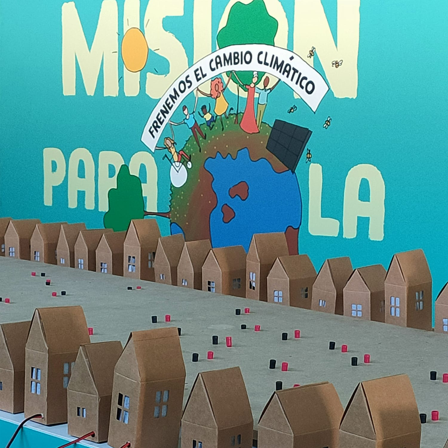 Vista lateral de un cartel que dice «Misión para la humanidad» y «Frenemos el cambio climático» junto a un dibujo del planeta tierra y figuras humanas. Frente a él, una mesa con maquetas de casas de cartón, cables eléctricos y zonas verdes.