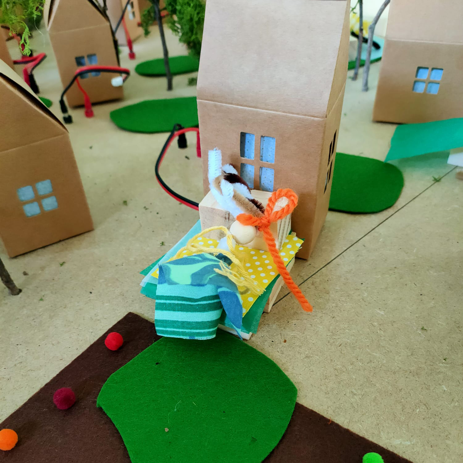 Vista detalle. En el centro, un muñeco de trapo con figura humana frente a una casa de cartón, que es parte de una comunidad de casas de cartón, zonas verdes, árboles y cables eléctricos.
