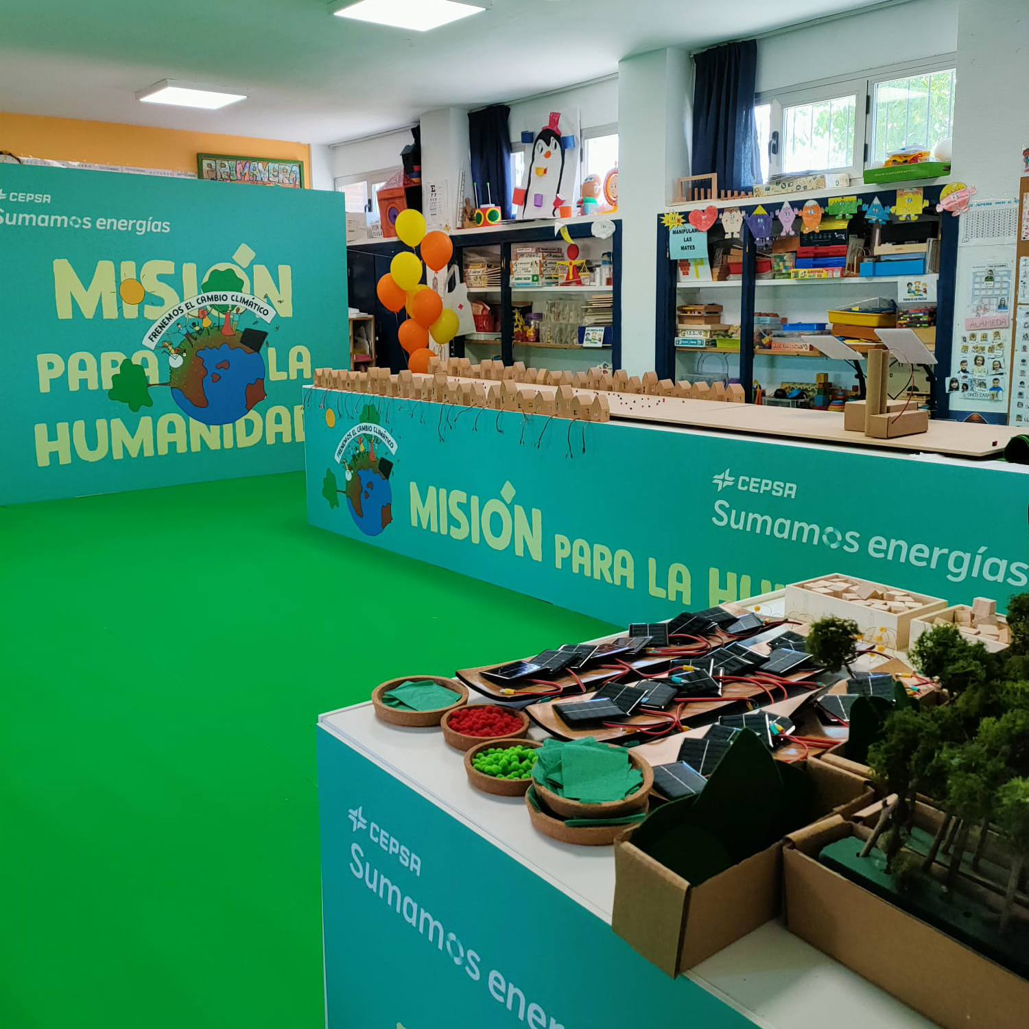 Vista general de la sala de exposición. La sala tiene un suelo de color verde y varios carteles verdes con los mensajes «Cepsa, sumamos energía», «Misión para la humanidad» y «Frenemos el cambio climático». En el espacio se distribuyen maquetas interactivas que incluyen casas de cartón, conexiones eléctricas, paneles solares y zonas verdes. Al fondo, una estantería repleta de materiales no relacionados con la exposición, como libros, juegos, manualidades y globos.