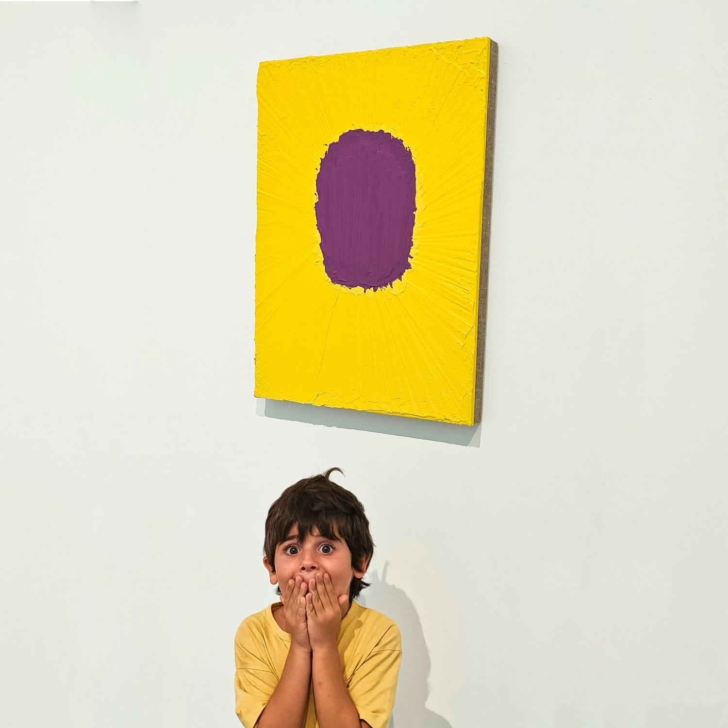 Vista frontal de un niño mirando a cámara mientras se tapa la boca con las manos y abre los ojos en señal de sorpresa. Tras él, la obra 'Incluso en la nieve de azufre', de fondo amarillo y en el centro una mancha de color violeta.