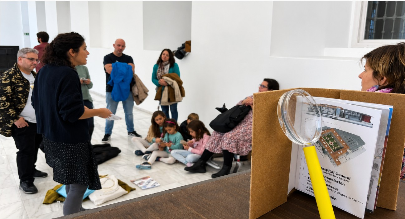 fotografia durante sesión Reina. En primer plano un cuadernito con imagen del Reina y lupa y al fondo familias escuchando