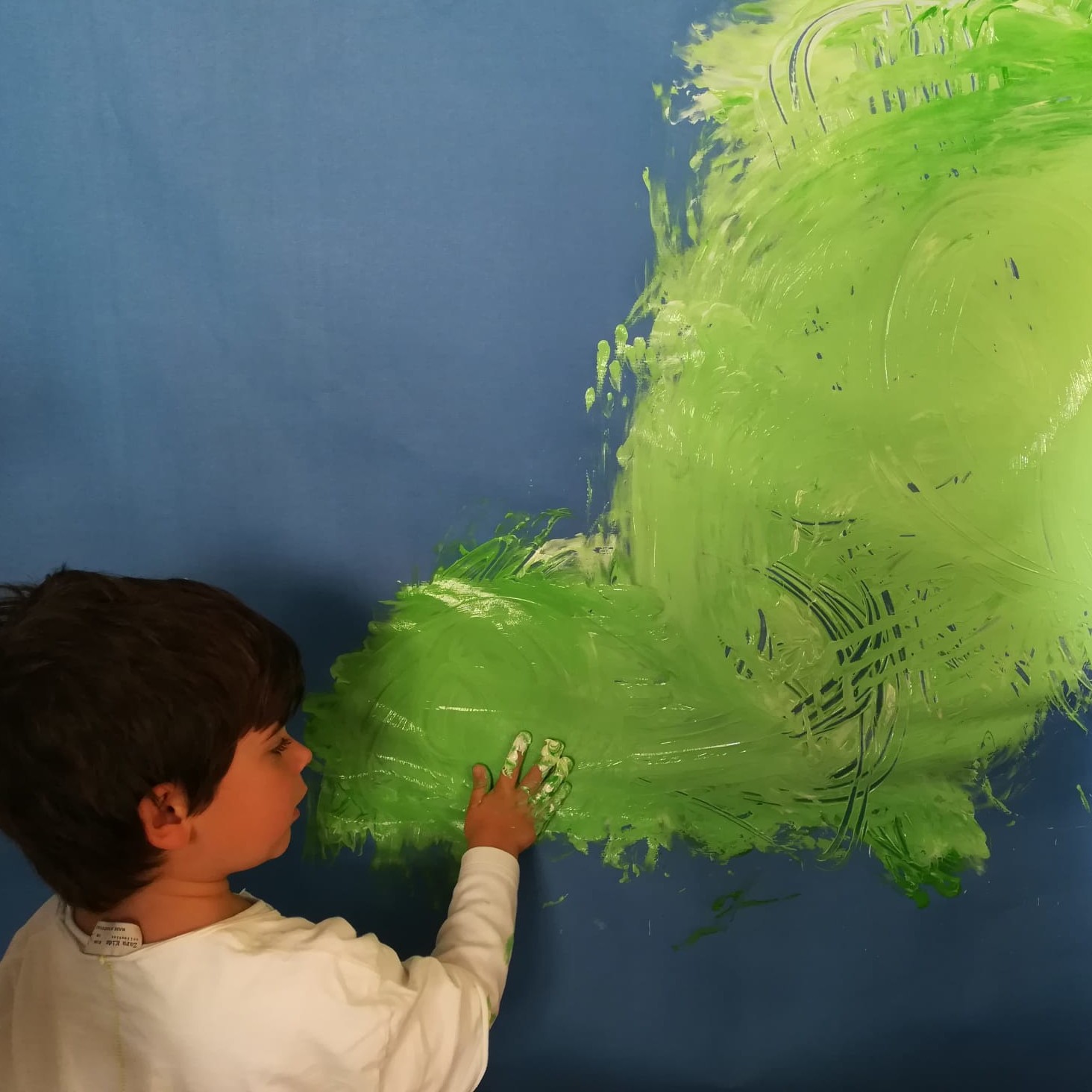 Vista posterior de un niño pintando con su manos trazos verdes sobre papel un papel azul en una Propuesta de experimentación de la obra The River.    https://cazapeonzas.com