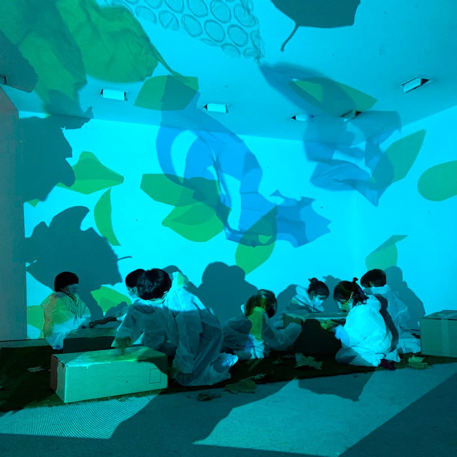 Vista lateral de siete niños, sentados en una habitación con imágenes proyectadas en las paredes, pintando cajas concentrados en el suelo.