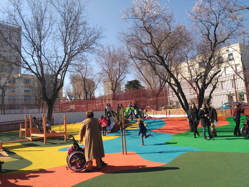 Parque Infantil Escuelas Bosque. [Descripción de imagen] Niños y niñas jugando en diferentes juegos. En primer plano, una niña en silla de ruedas. En el fondo, árboles y edificios.